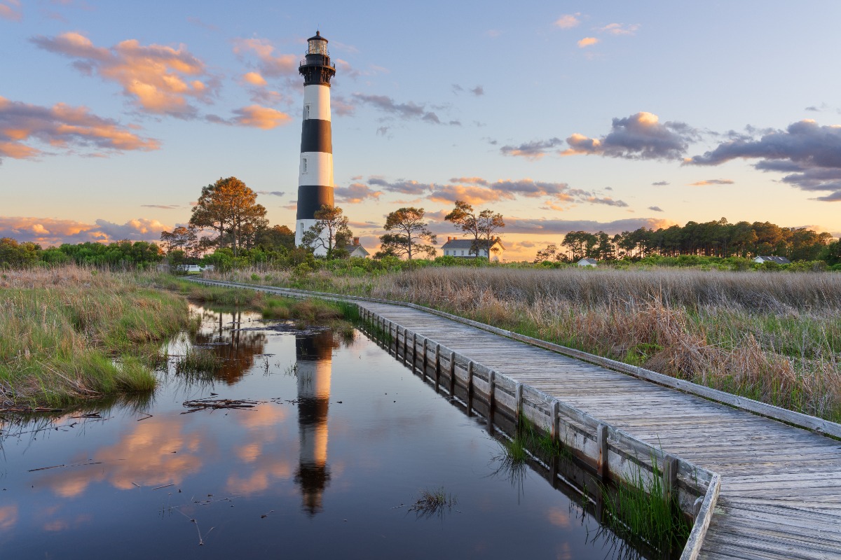 A striped lighthouse stands near a wooden boardwalk over calm marsh water at sunset, reflecting in the pond nearby.