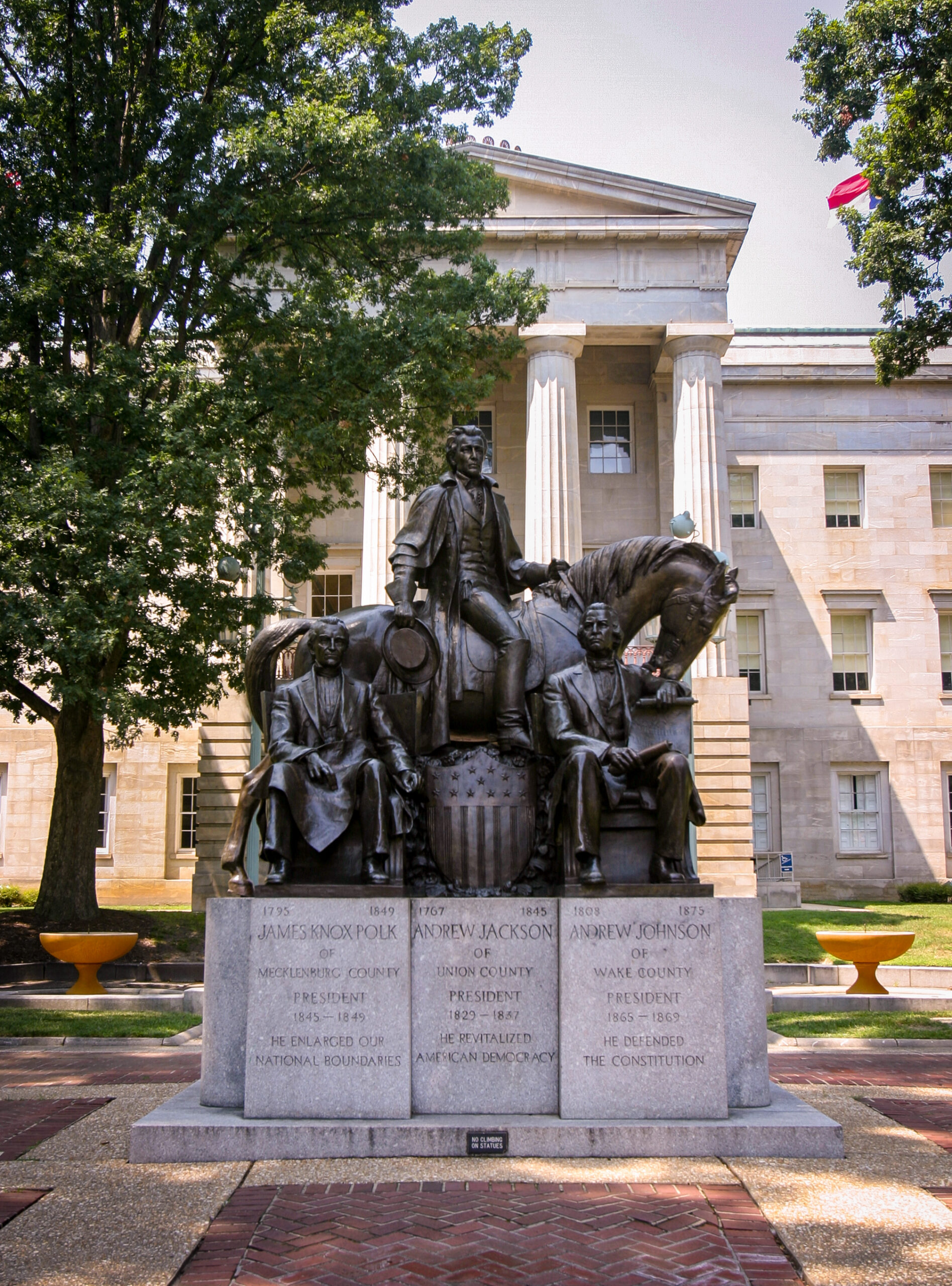 Monument with a bronze statue of three seated figures in front of a classical building with tall columns and a flag in the background, commemorating U.S. presidents James K. Polk, Andrew Jackson, and Andrew Johnson.