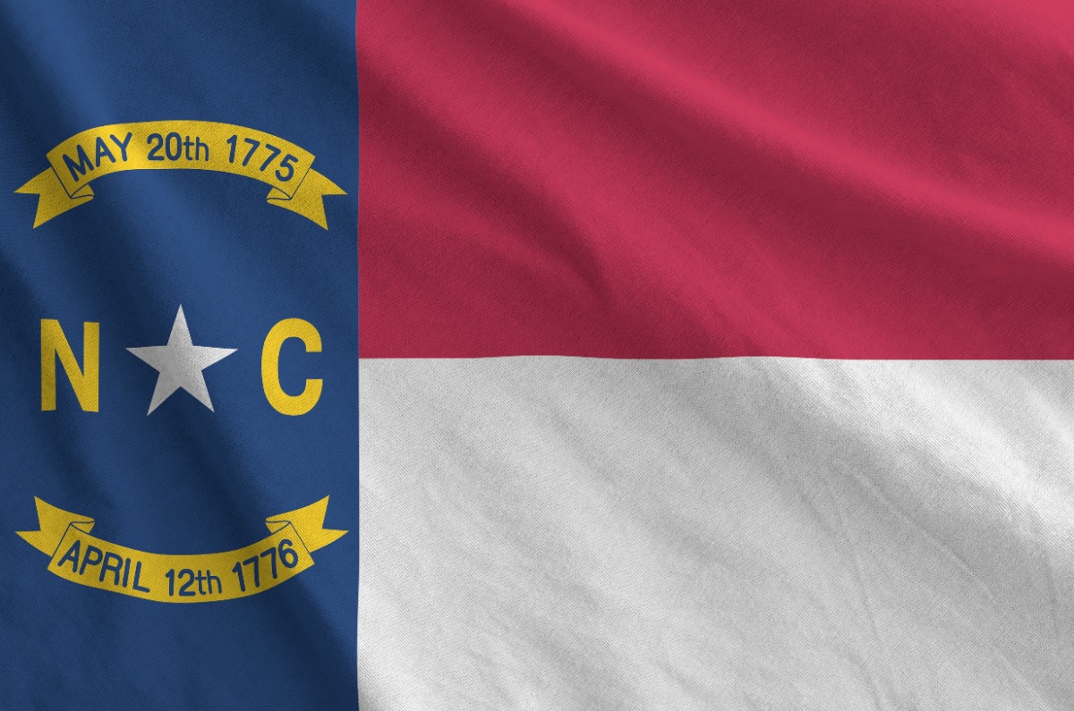 North Carolina state flag waving: blue vertical stripe with yellow 'N C' and white star, and two yellow ribbons reading 'May 20th 1775' and 'April 12th 1776' on a red and white field.
