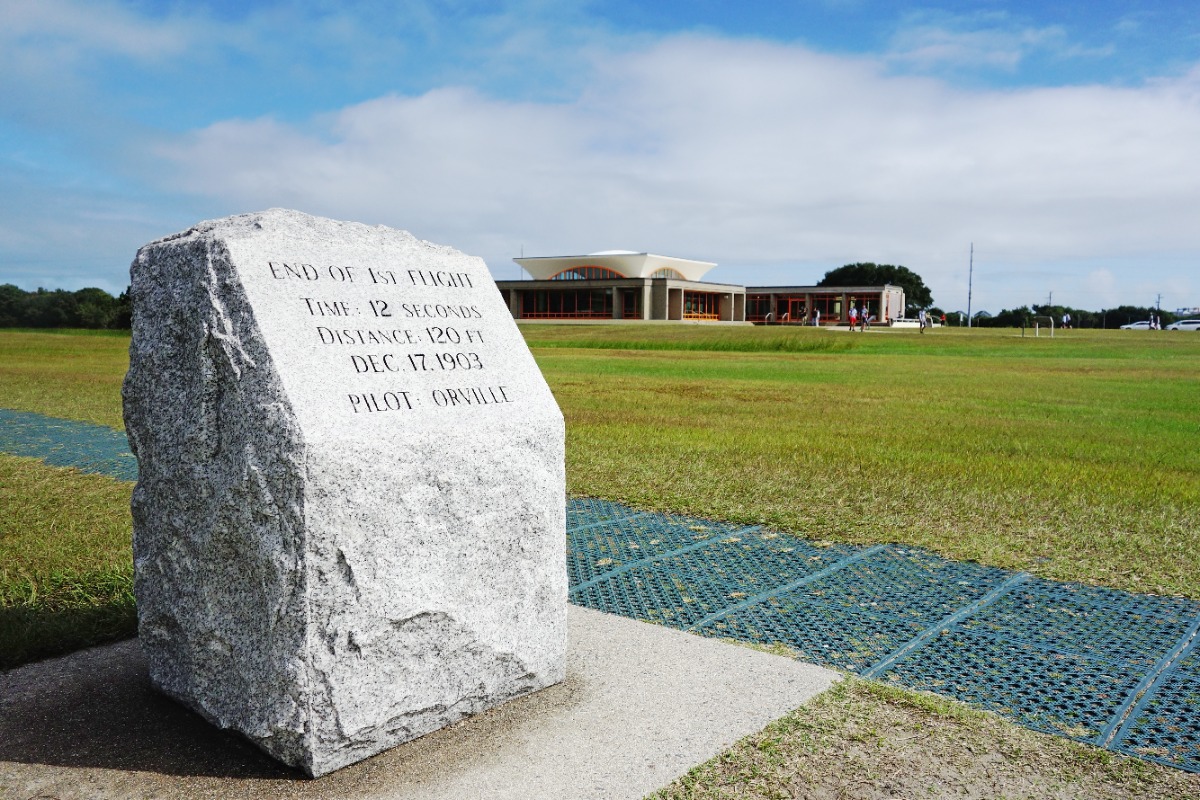 Stone monument with engraved text commemorating the first flight (Orville Wright, 1903) at a grassy airfield with a modern building in the background.