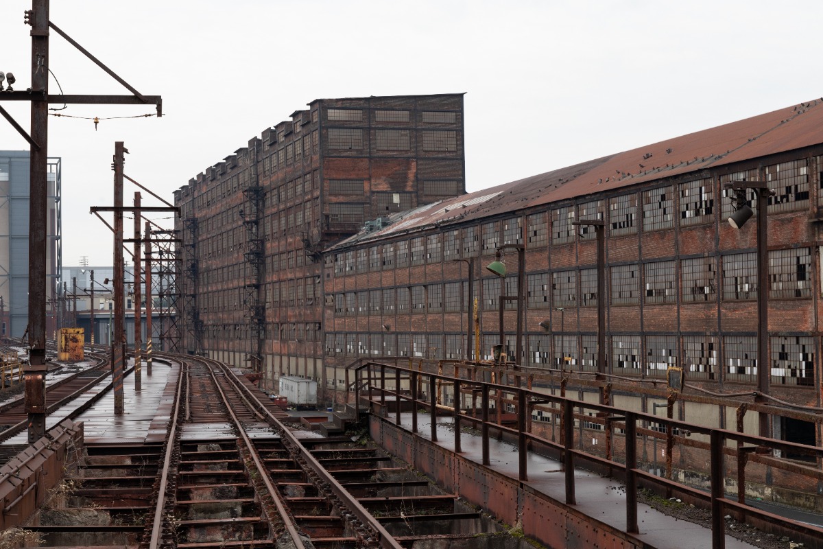 Abandoned industrial complex with rusted rail tracks beside a long brick warehouse under an overcast sky.
