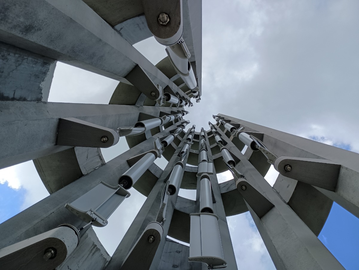 Looking up at a spiraling steel sculpture with cylindrical tubes attached to concrete supports against a cloudy sky.