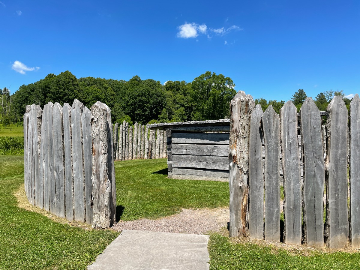 A circular enclosure of tall weathered wooden stakes surrounds a small, gray log shed on a grassy field under a bright blue sky.