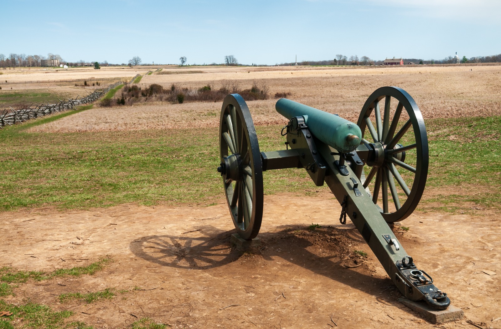 Green field cannon with large wooden wheels set on a dirt patch in an open rural landscape.