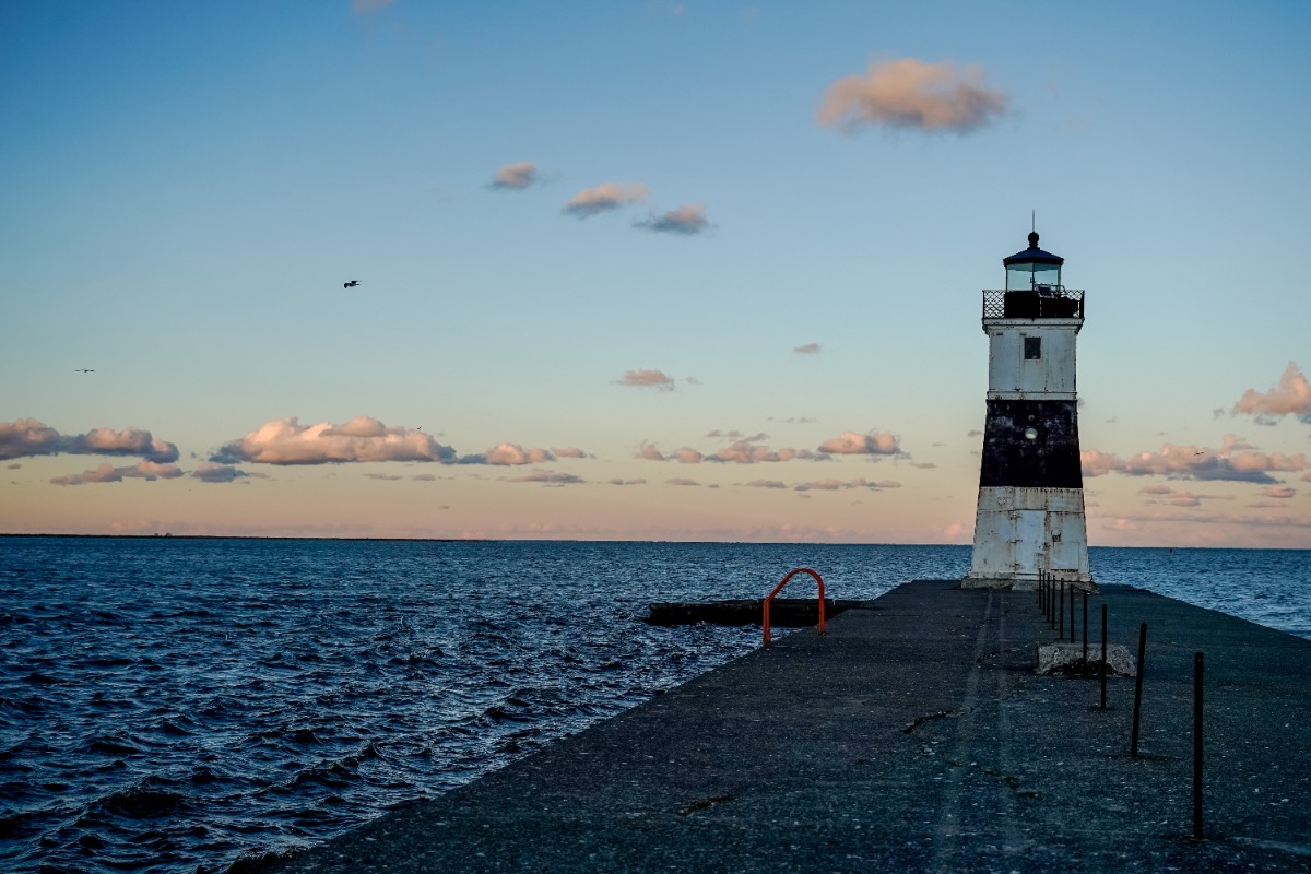 Lighthouse at the end of a concrete pier extending into a calm sea at sunset with blue sky and scattered clouds