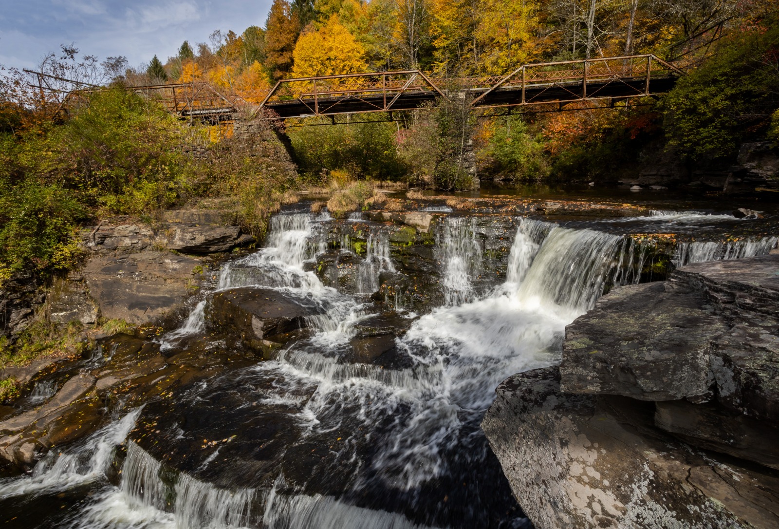 Waterfall cascading over dark rocks beneath a rusted metal bridge, surrounded by autumn-colored trees.