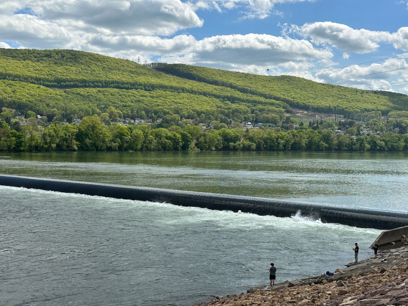 Wide river with a low dam, tree-lined shore and green hills in the distance; a few people fish on the rocky bank of the river.