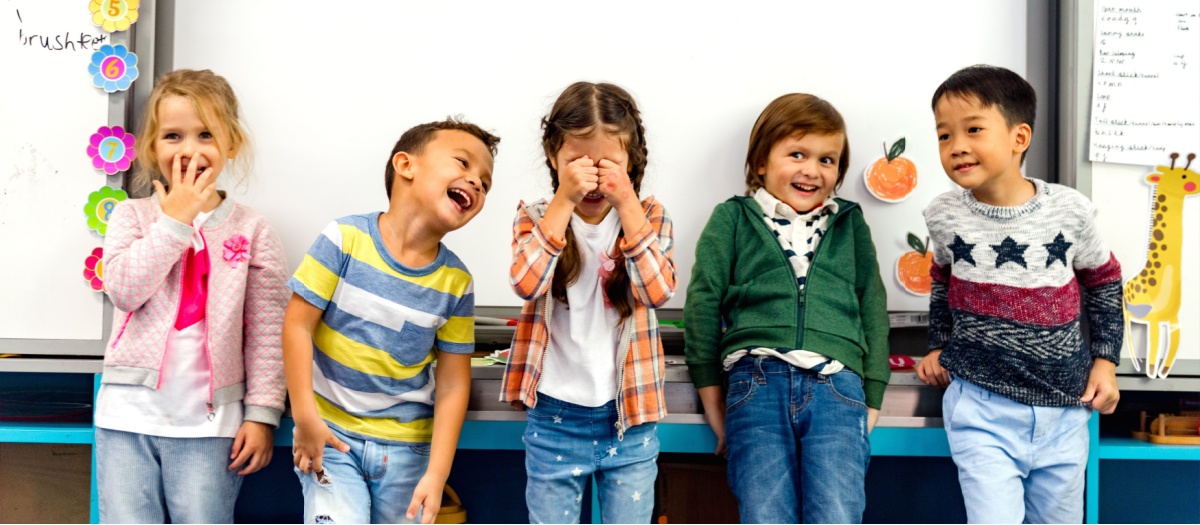 Five happy children stand in a bright classroom, with one girl covering her eyes as others smile nearby, colorful decorations on the wall behind them.