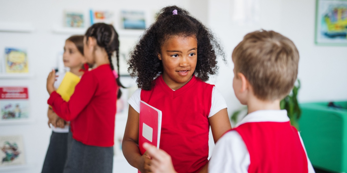 Two children in red uniforms talk in a bright classroom, one holding a red notebook.