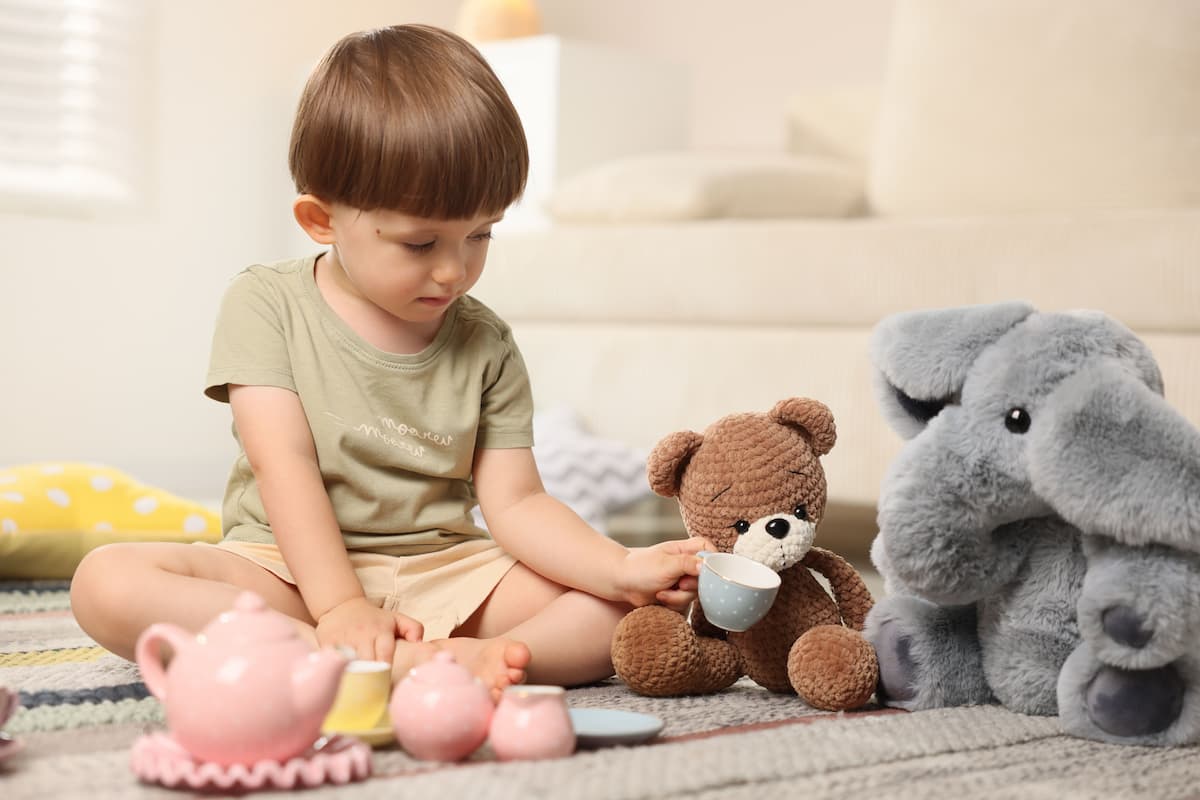 Toddler sits on a rug, pouring pretend tea for a brown teddy bear and a gray plush elephant with a small tea set.