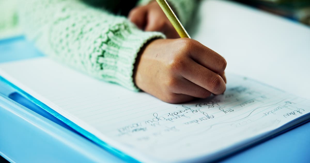 Close-up of a young student’s hand writing in a lined notebook with a yellow pencil, wearing a mint-green sweater sleeve.