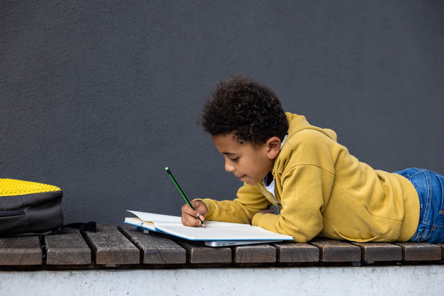 Boy lying on his stomach on a wooden bench, writing in a notebook with a green pencil, a yellow hoodie and jeans, a backpack beside him after-school scene.