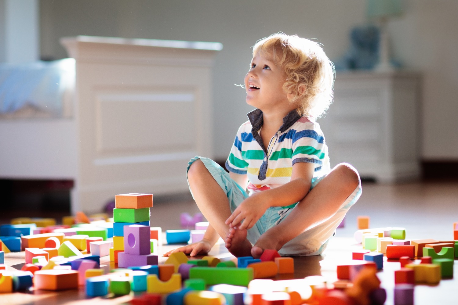 Young child sits cross-legged on a sunlit floor, smiling while playing with colorful wooden blocks scattered around.