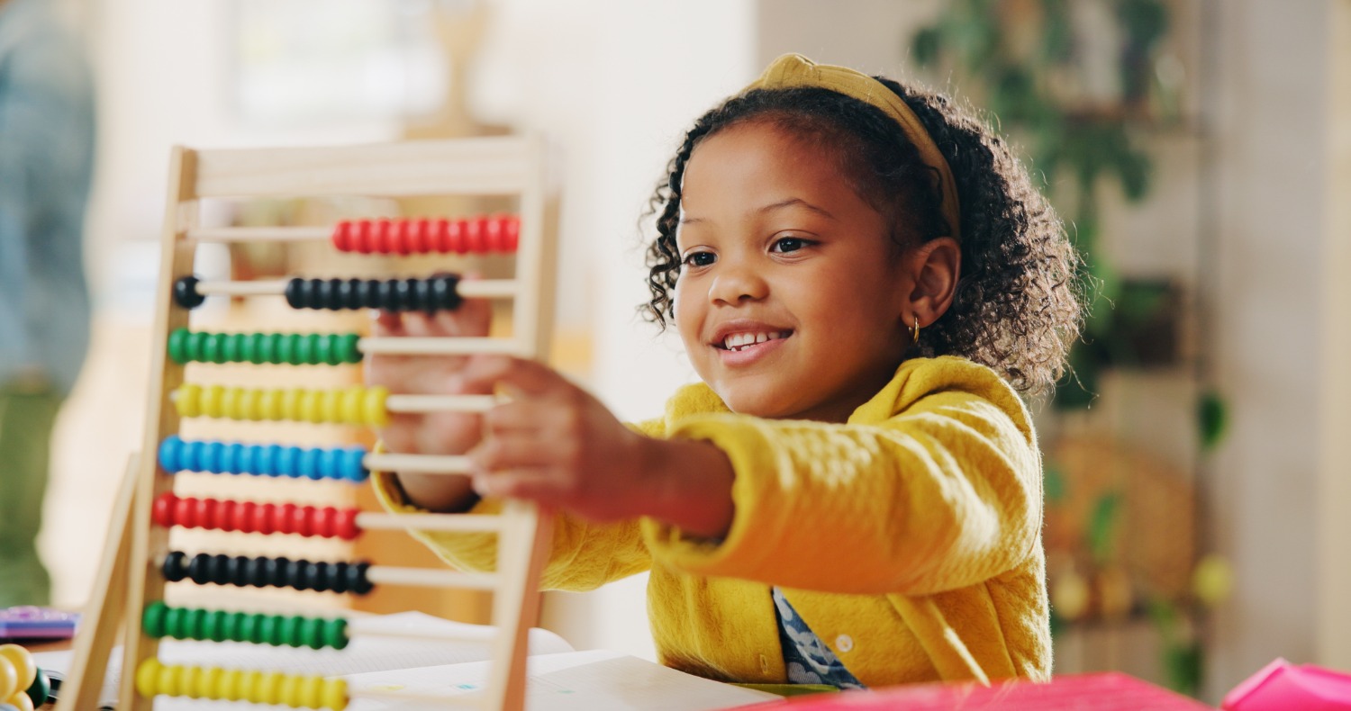 Girl with a yellow sweater playing with a wooden abacus, smiling as she counts beads in a bright room.