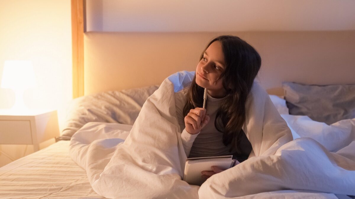 Young girl in a white duvet, leaning in bed and writing in a notebook with a pen, illuminated by a warm bedside lamp.