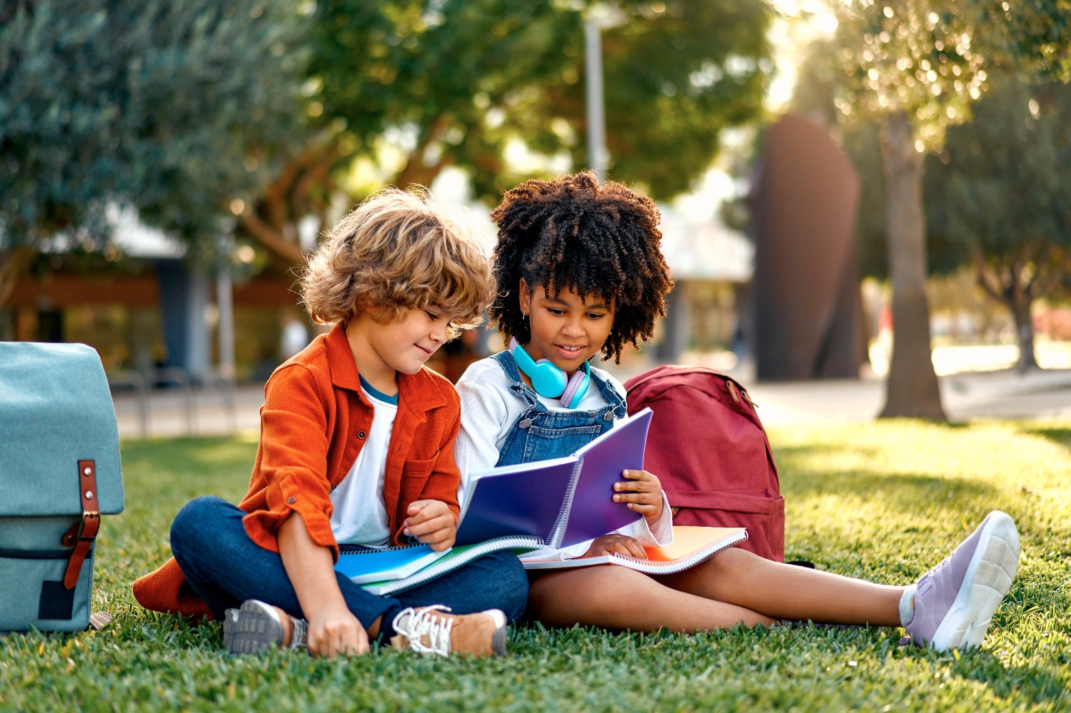 Two kids sit on grass reading notebooks together with backpacks nearby in a sunny park.