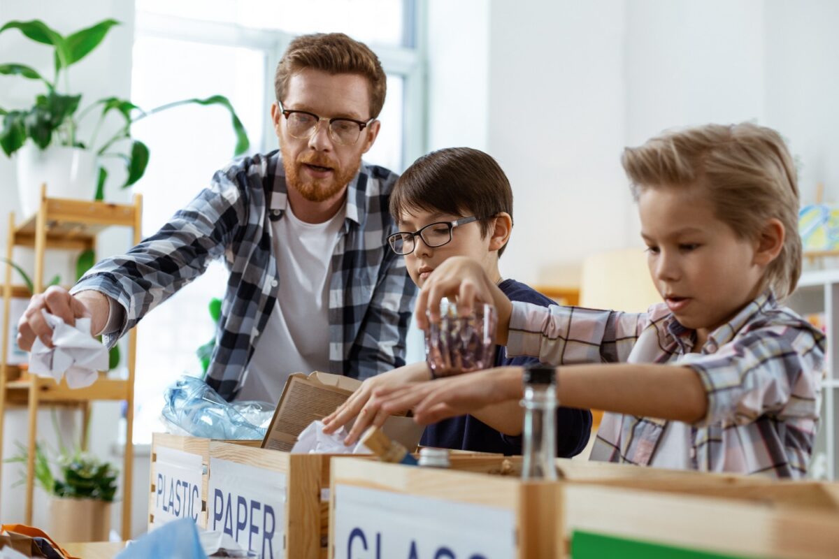 Man and two boys sorting recyclables into labeled bins in a bright room.