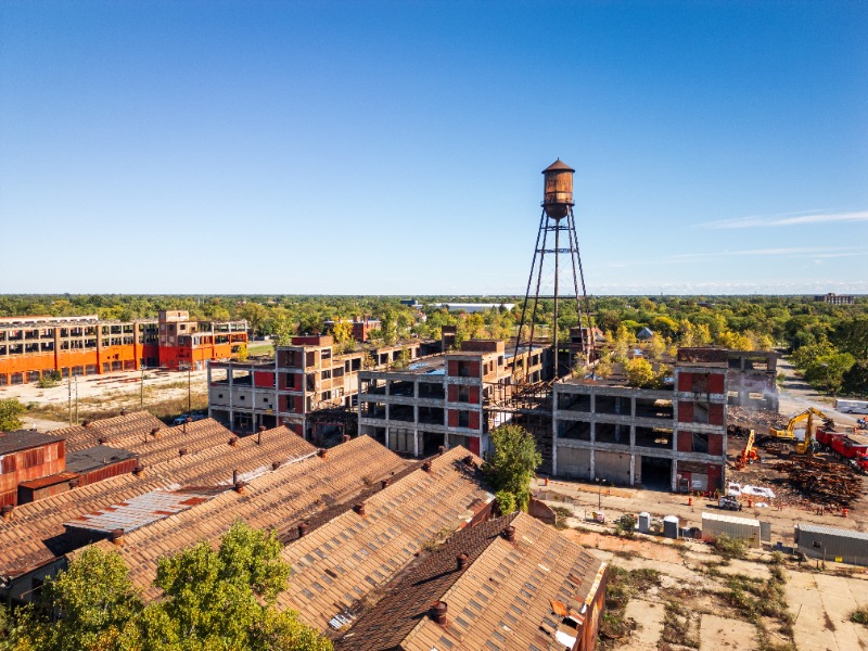 Aerial view of an abandoned industrial complex with dilapidated brick buildings and a tall rusted water tower under a clear blue sky, trees in the distance.