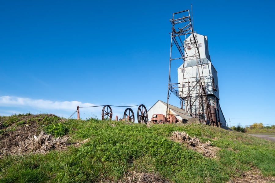 Abandoned mining headframe and rusted gear machinery on a grassy hillside under a bright blue sky, daylight.