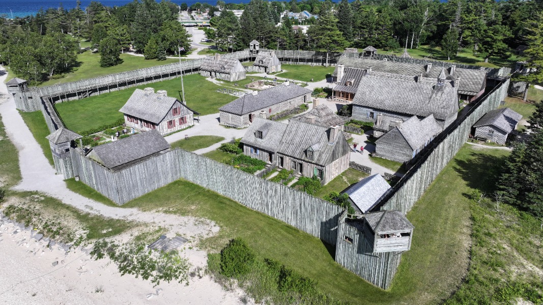 Aerial view of a wooden palisaded fort with watchtowers and clustered buildings inside, set on green grass near a forest and lake area.