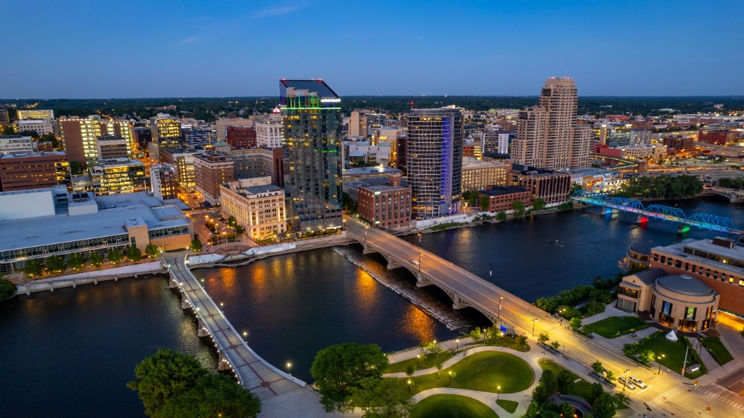 Aerial view of a city riverfront at dusk with lit buildings, two bridges crossing the water, and a curved riverside park in the foreground.