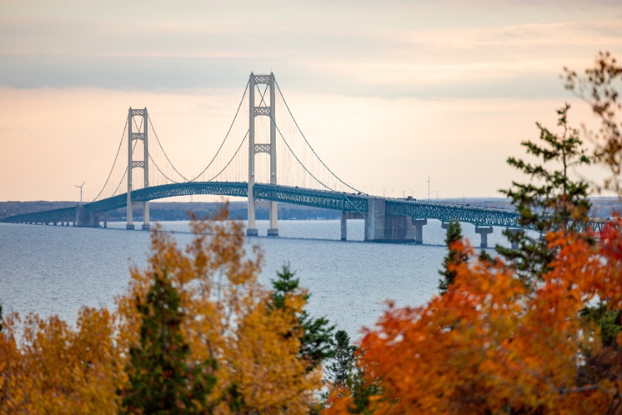 Suspension bridge spanning a wide body of water with autumn trees in the foreground and a soft sunset sky behind it