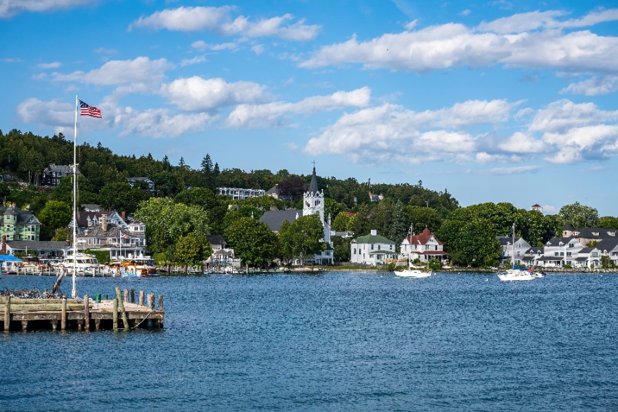 Lakeside town with a wooden pier, American flag, boats on the water, and a white church tower among trees.