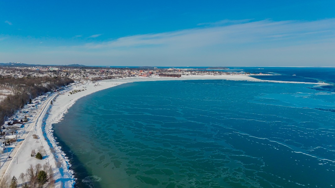 Aerial view of a snowy coastal town with a curved blue bay and partially frozen water, buildings visible in the distance.