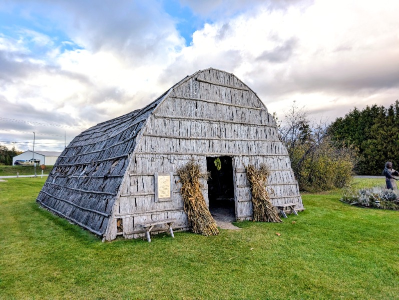 Curved, weathered wooden hut on a green lawn, with bundles of dried cornstalks at the doorway.