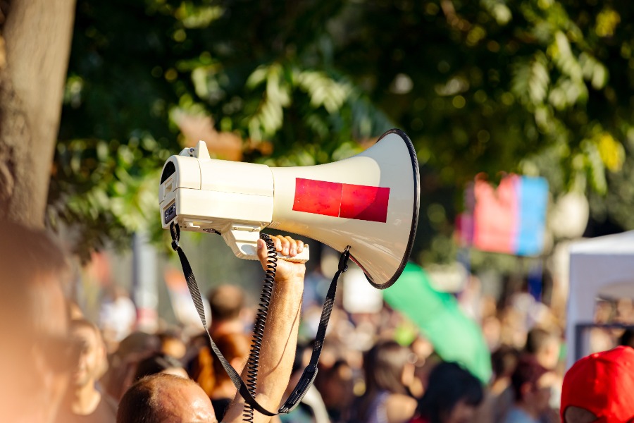 Person raising a megaphone above a crowded protest with colorful banners in the background.