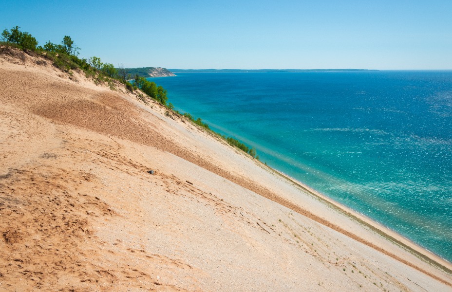 Slope of sandy dunes meeting bright blue sea under a clear sky; green shrubs line the crest on the left side. SE coastline view.