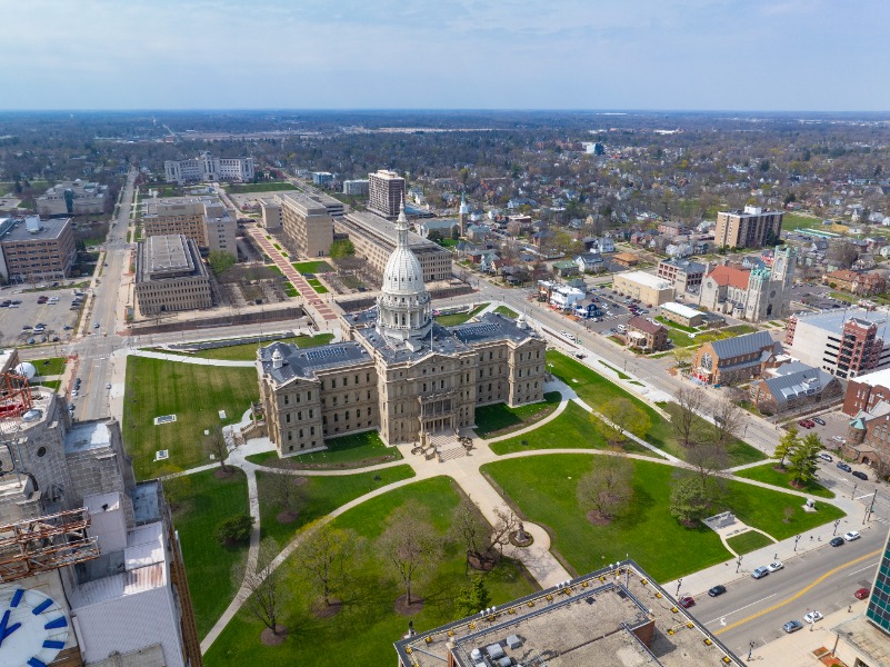 Aerial view of a domed government building surrounded by green lawns in a cityscape.
