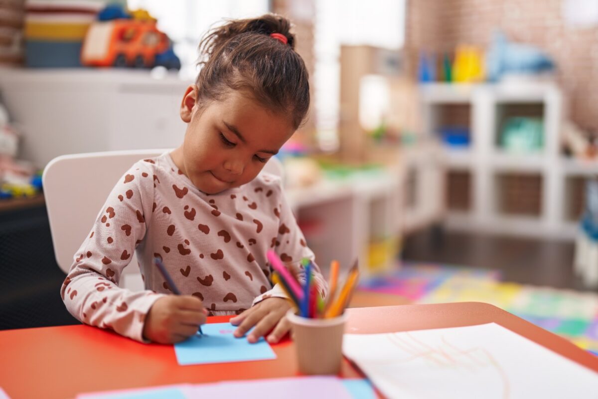 Young girl sits at a bright orange table, coloring on blue paper with colored pencils in a cup, in a cozy playroom.