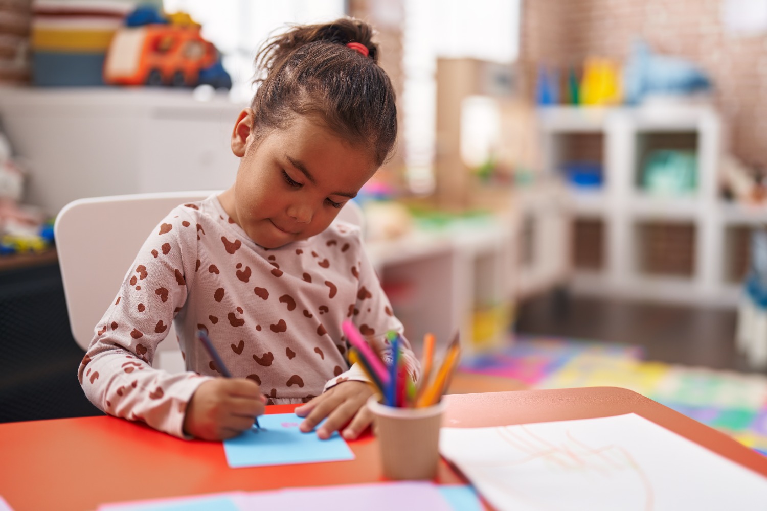 Young girl sits at a bright orange table, coloring on blue paper with colored pencils in a cup, in a cozy playroom.