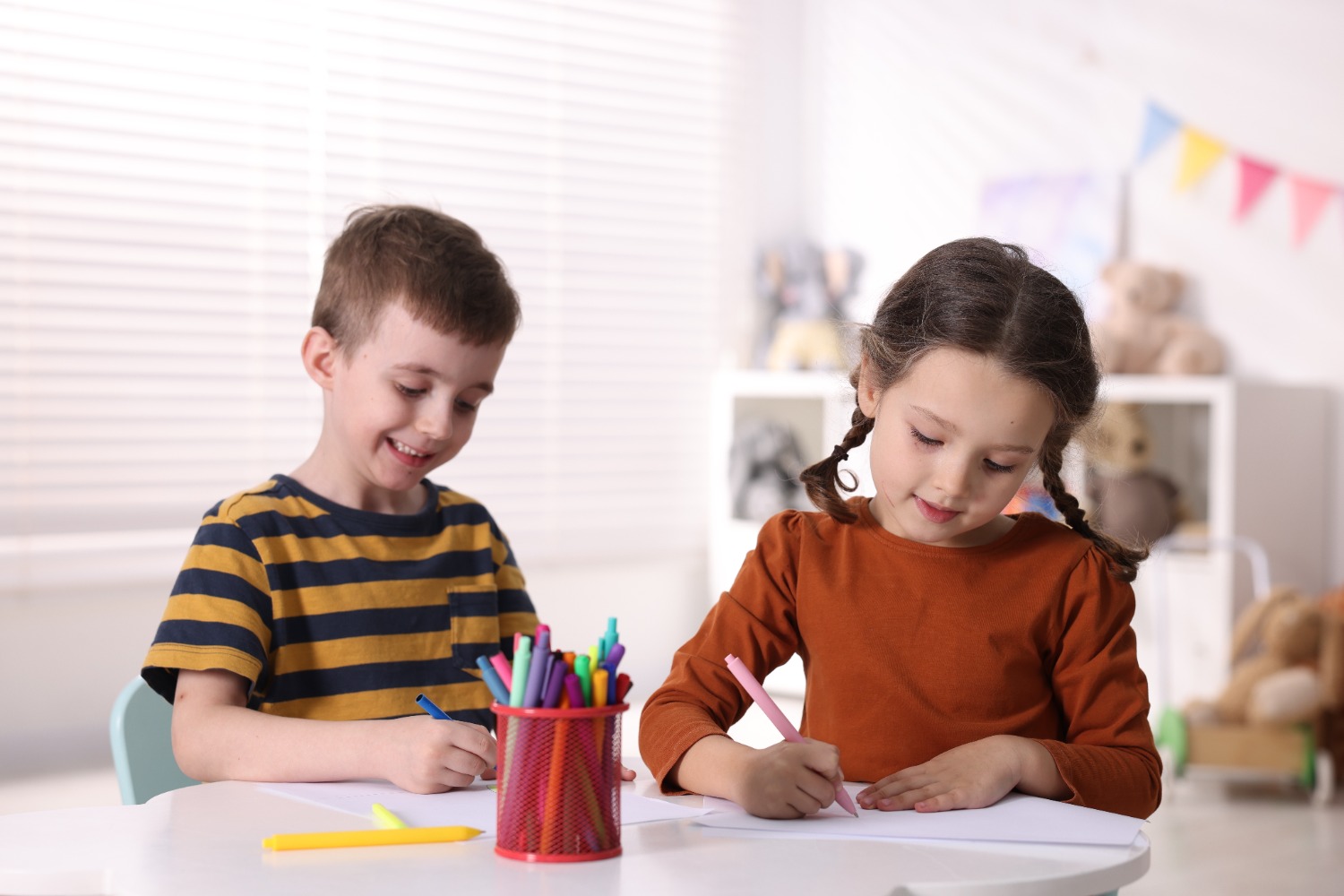 Two children, a boy and a girl, sit at a white table drawing with colored markers in a bright playroom.