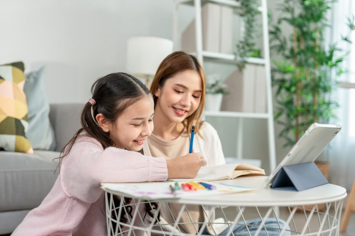 Mother and daughter studying together at a white table with a tablet, notebooks, and colored pencils in a bright, cozy living room