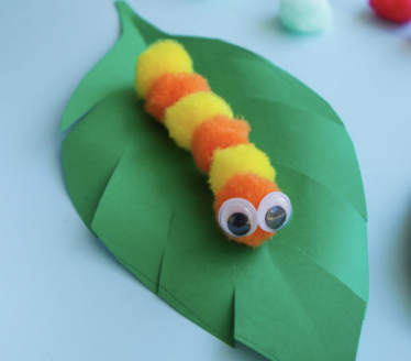 Yellow and orange pom pom caterpillar with googly eyes resting on a green paper leaf.