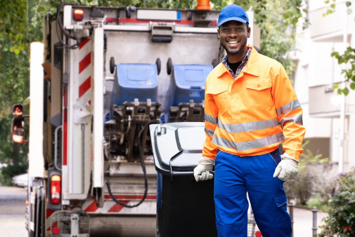 Smiling sanitation worker wearing an orange safety jacket and blue uniform standing beside a garbage truck while collecting trash.