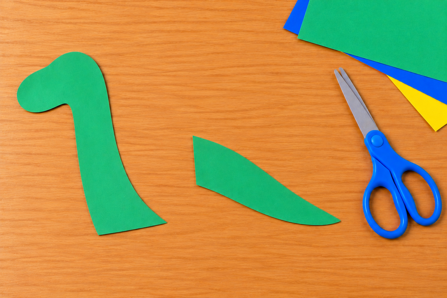 Green construction paper dinosaur head, long neck, and tail beside blue child-safe scissors.