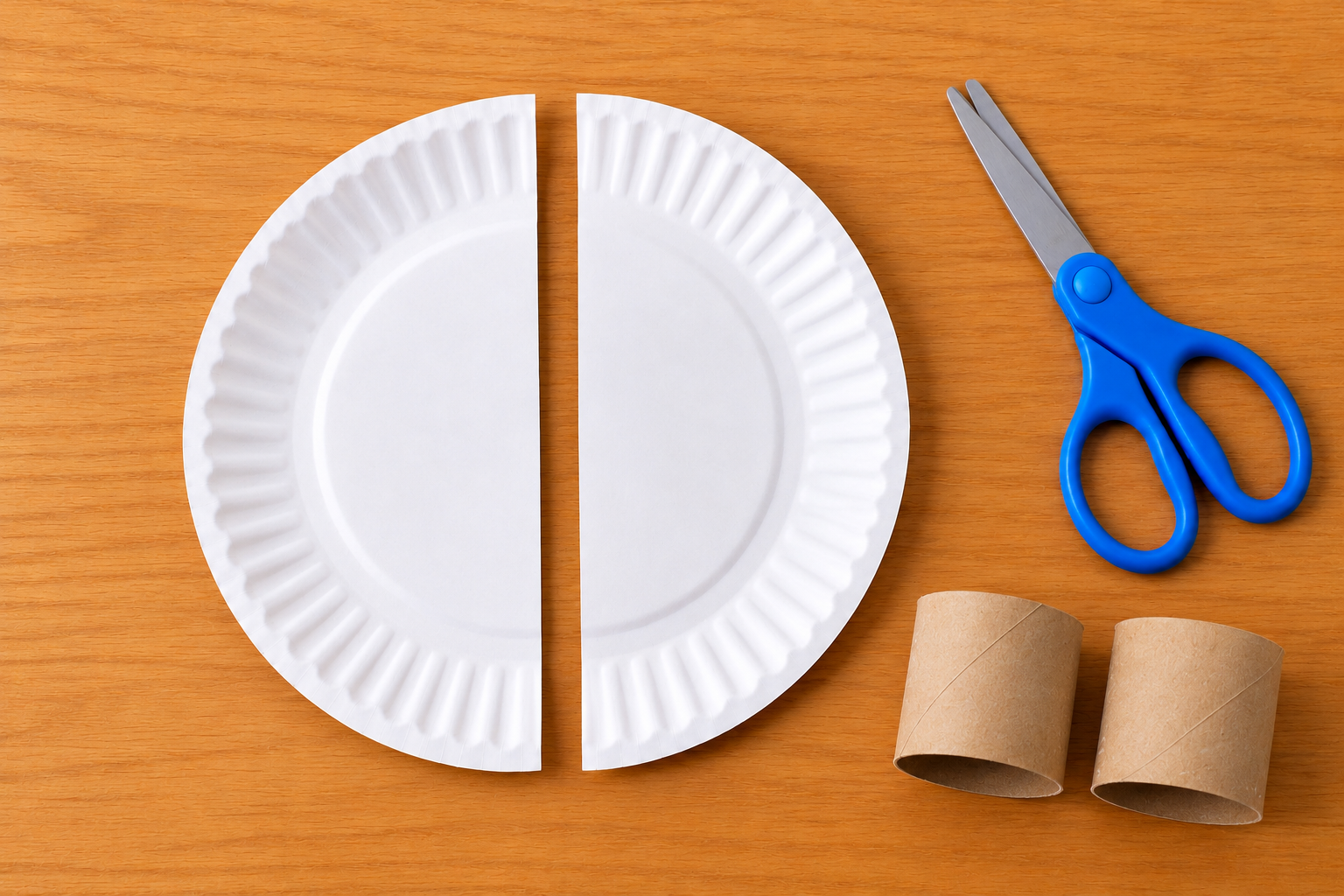 Paper plate cut in half beside blue scissors and a cardboard toilet paper roll cut into two pieces.